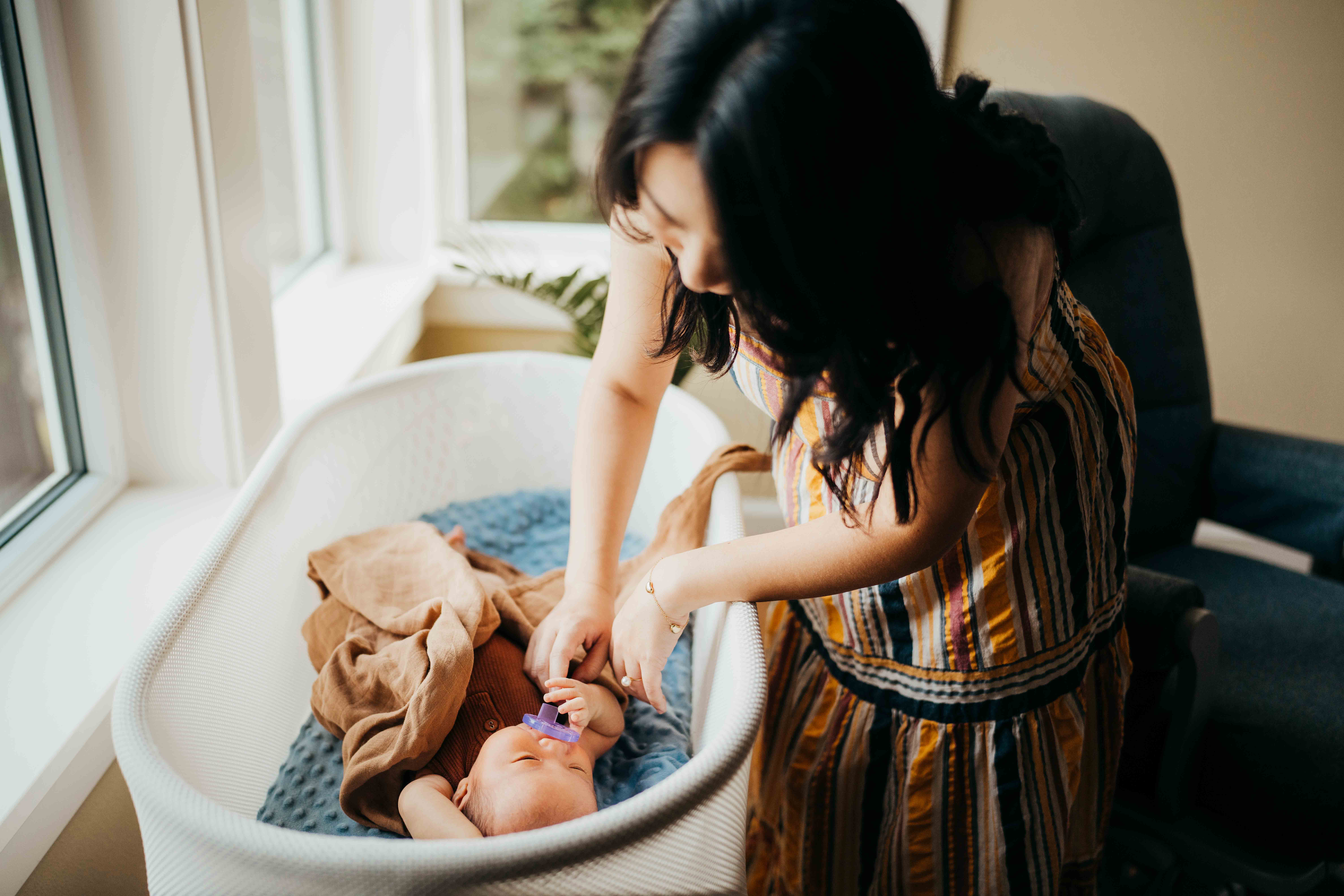 A new mom looks down on her newborn baby in a crib under a window in a stripe dress before getting pelvic floor therapy in seattle