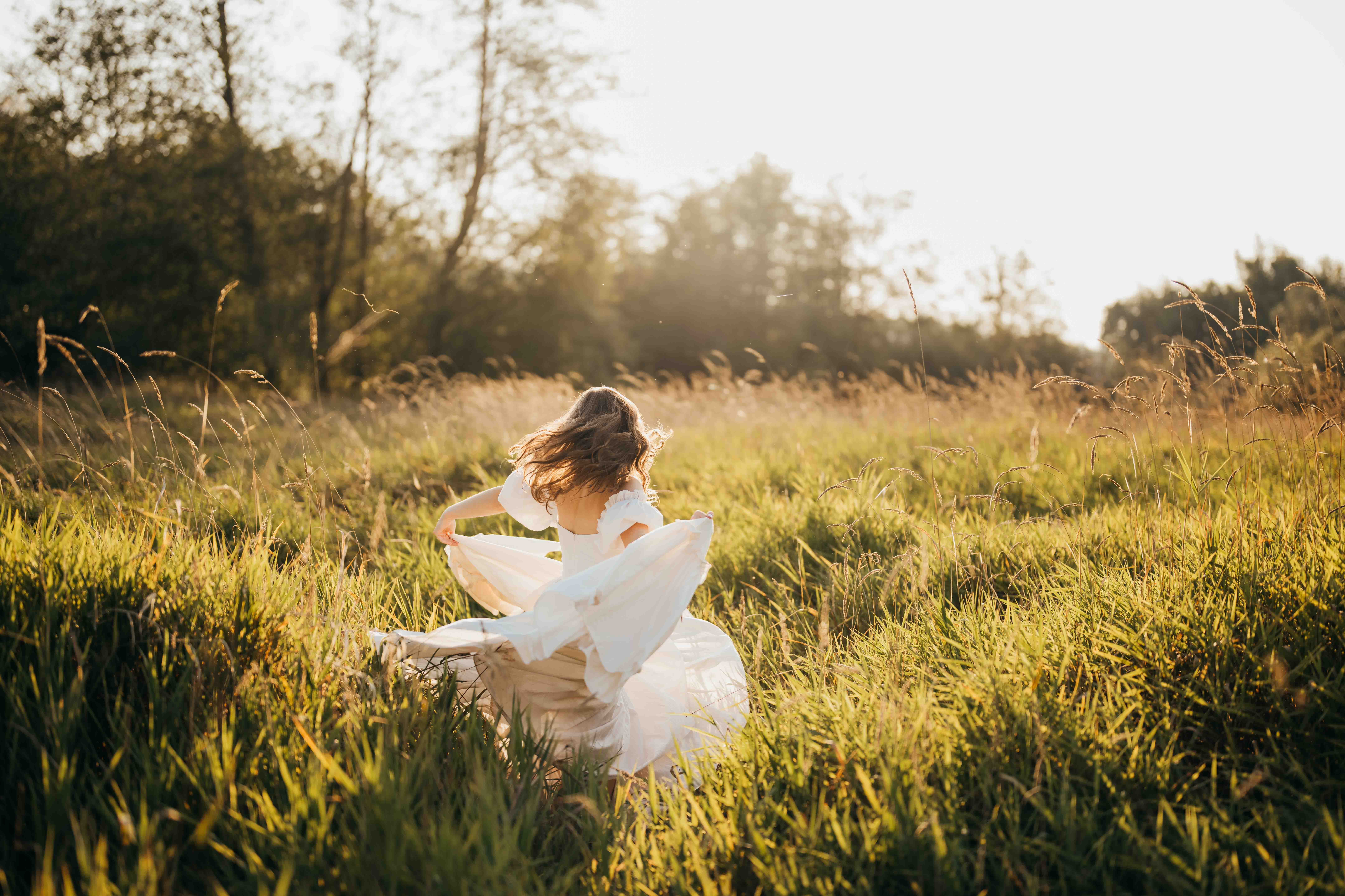 A woman dances in a white dress playing with the twirling train in tall grass at sunset