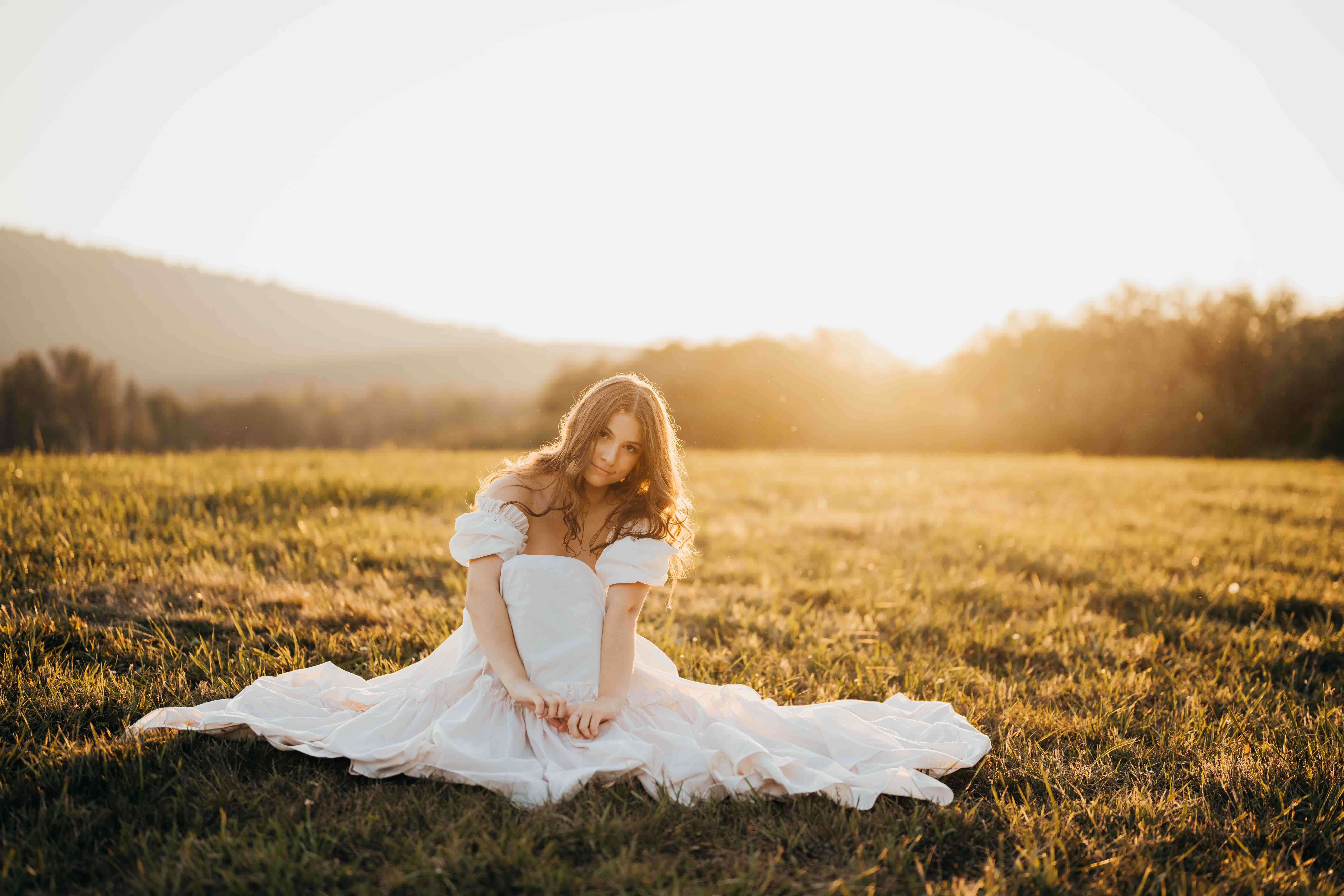 A high school senior sits in a meadow in a white dress at sunset after visiting tanning salons in seattle