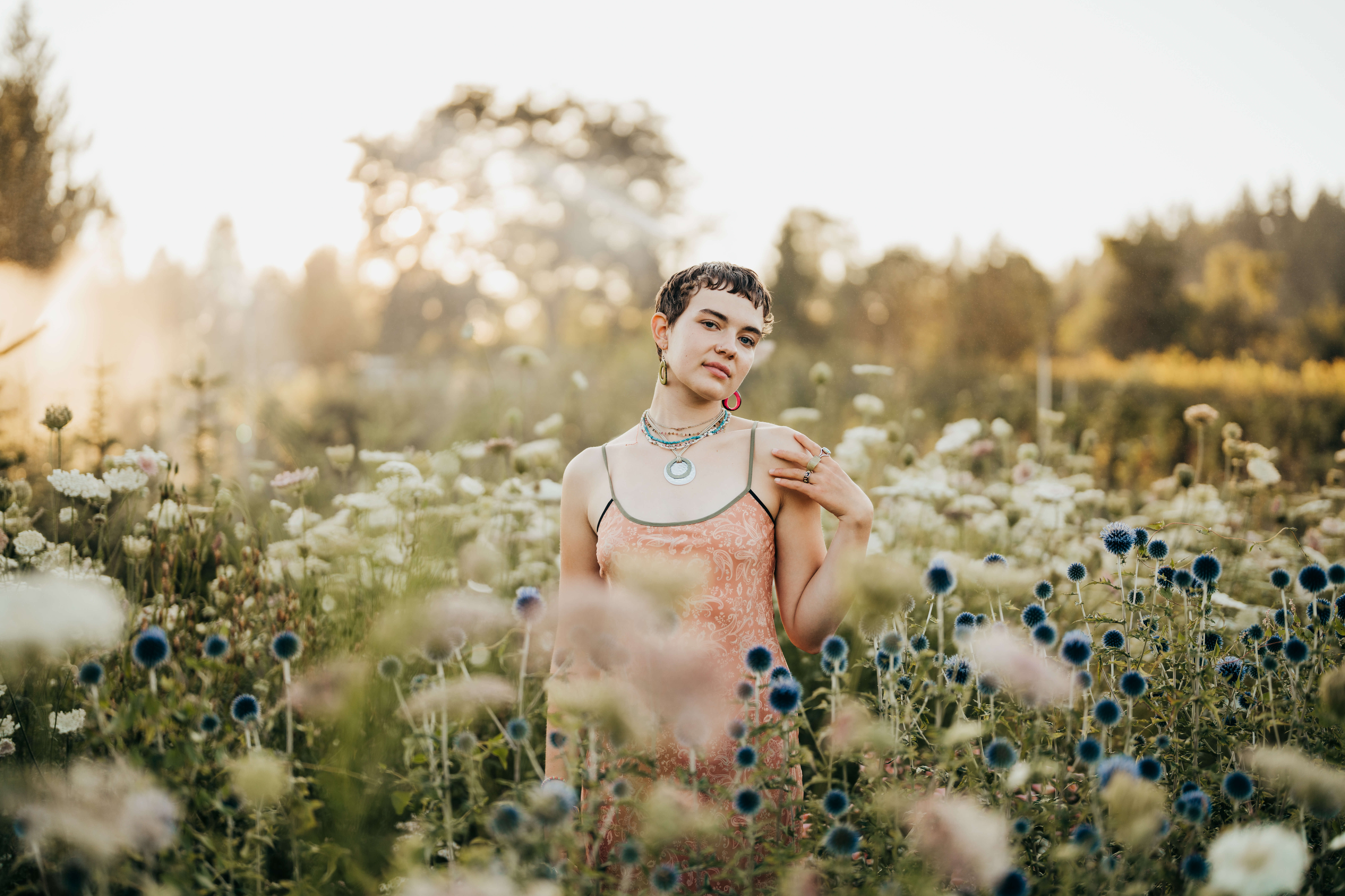 A high school senior in a pink dress stands among tall white and blue wildflowers at sunset in a garden