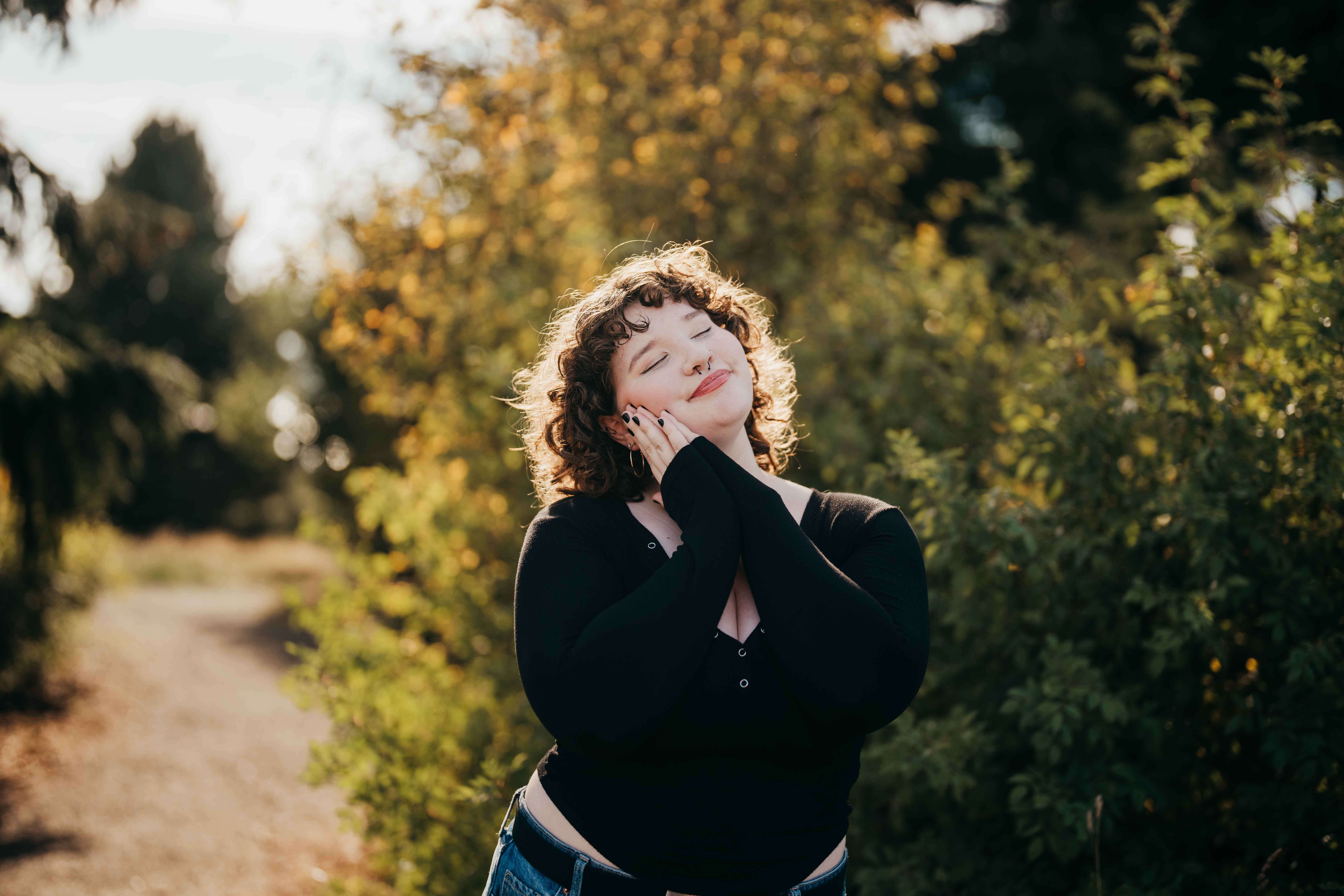 A high school senior in a black blouse smiles with hands on her cheek in a garden at sunset after using College in The High School