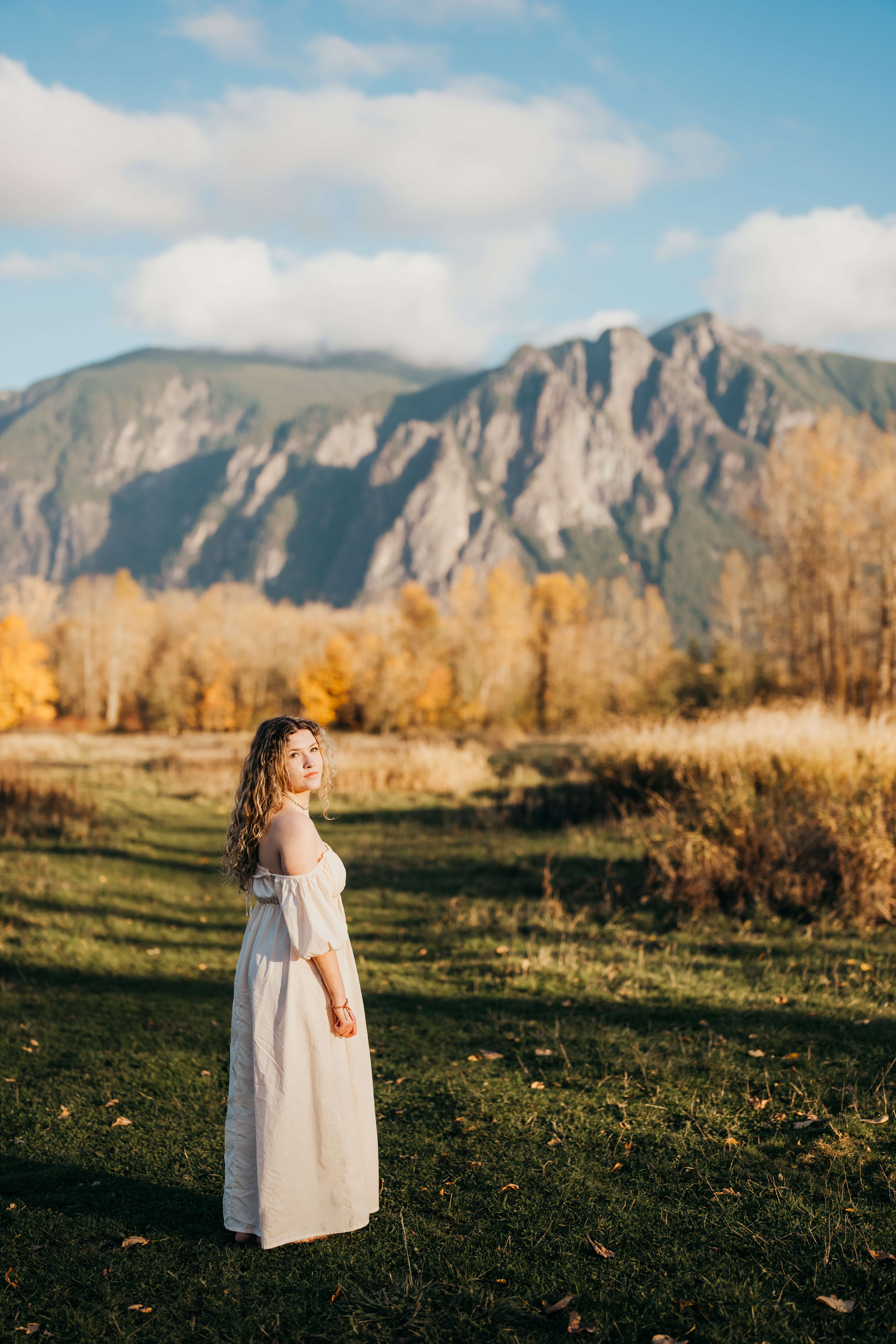 A high school grad looks back over her shoulder while walking a meadow by the mountains at sunset