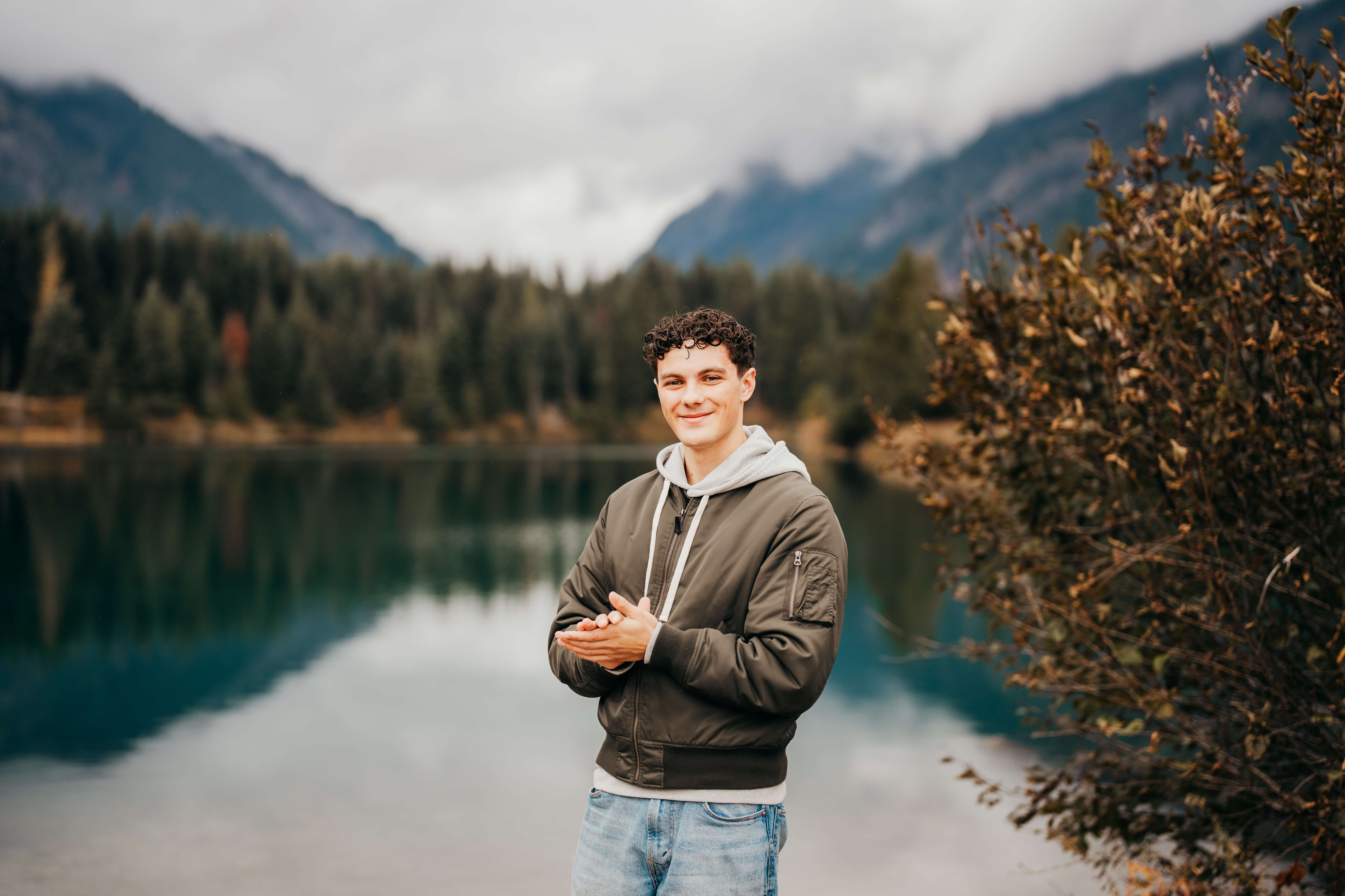A high school senior in a black bomber jacket and jeans stands by an epic lake smiling