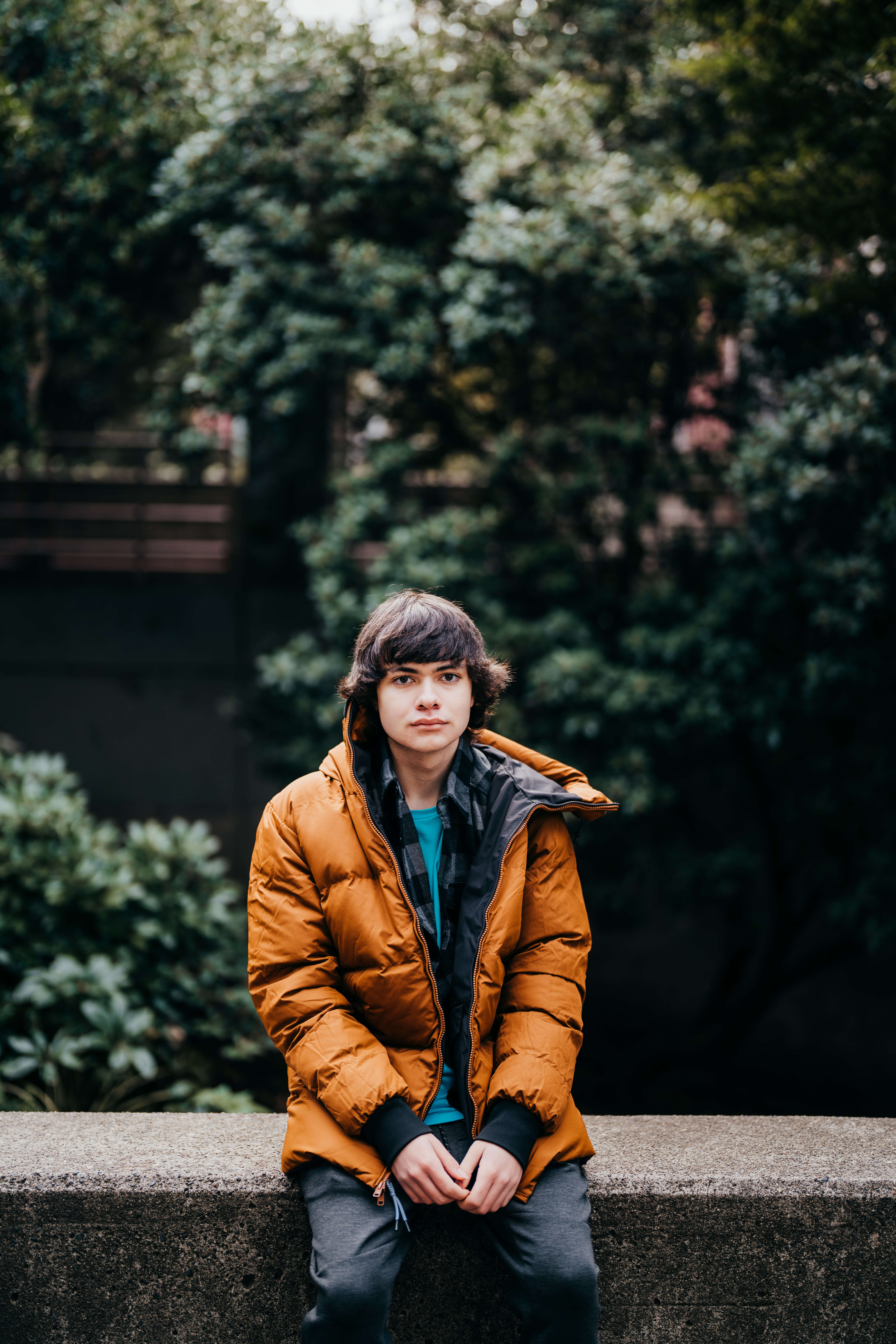 A teenager in an orange coat sits on a concrete railing in a park