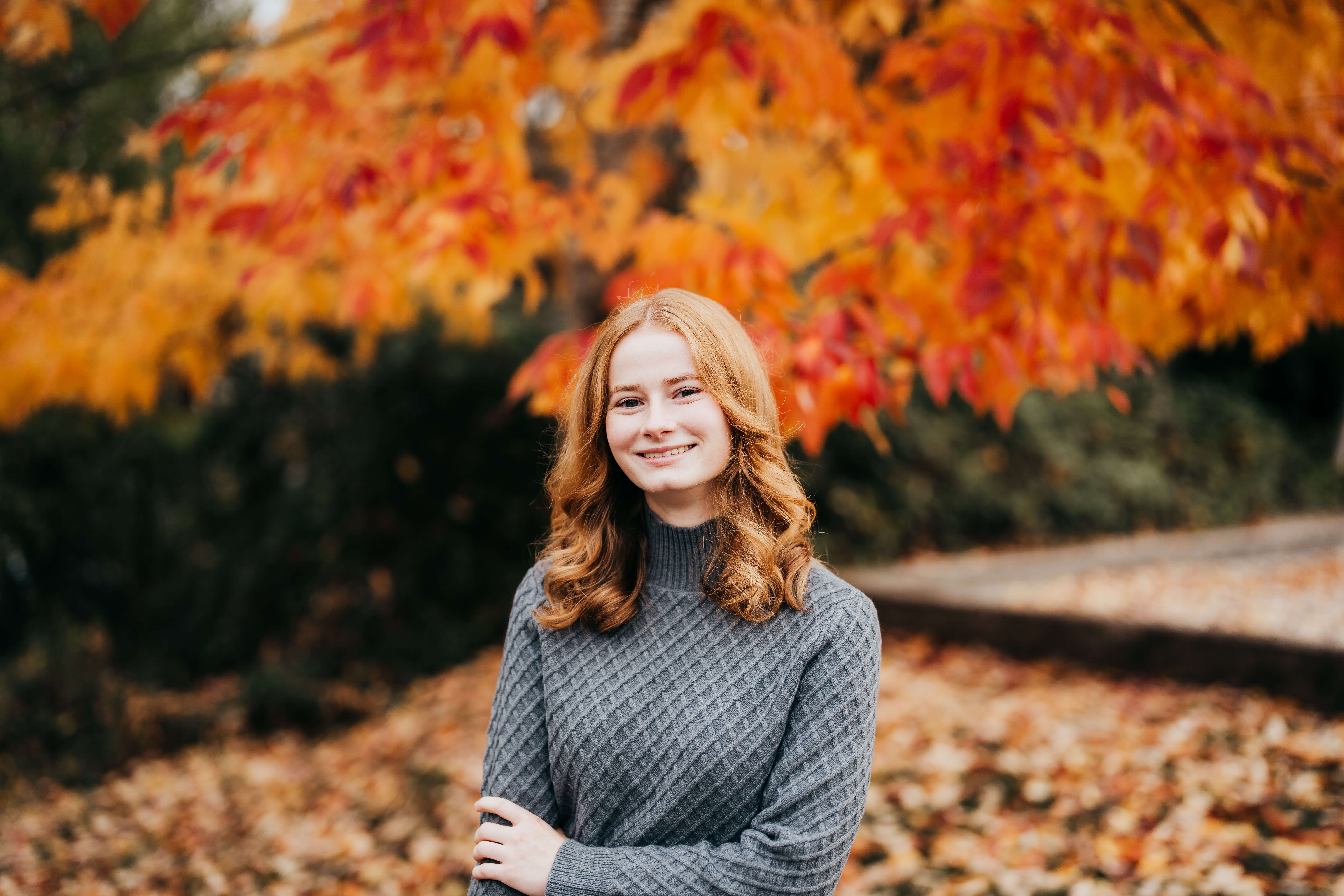 A high school senior with red hair stands under a fall colored tree in a grey sweater after visiting hair salons in seattle