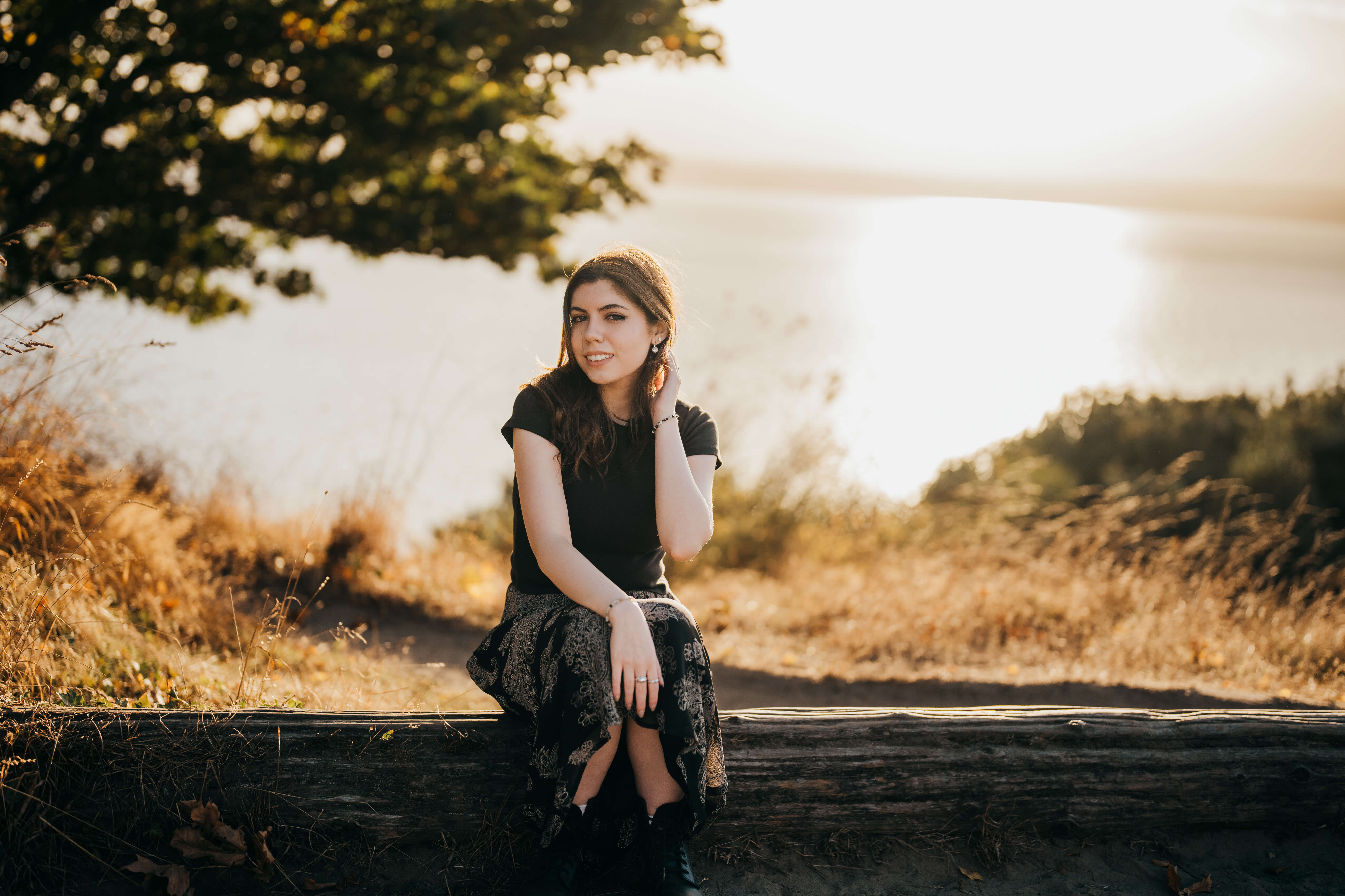 A high school senior in a black skirt and blouse sits on a log overlooking the water at sunset with a hand in her hair after visiting nail salons in seattle