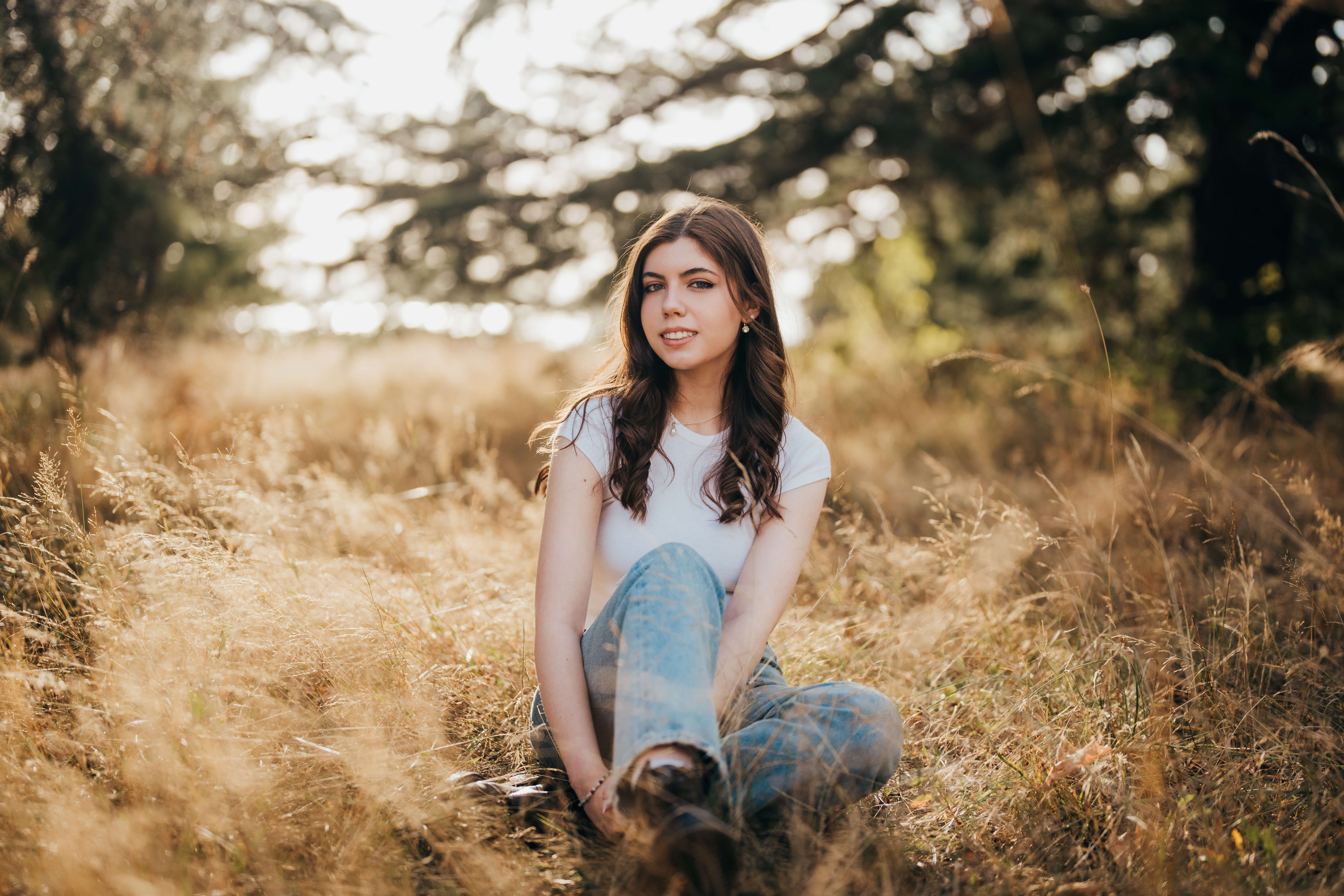 A high school senior in jeans and white blouse sits in tall golden grass smiling after visiting nail salons in seattle
