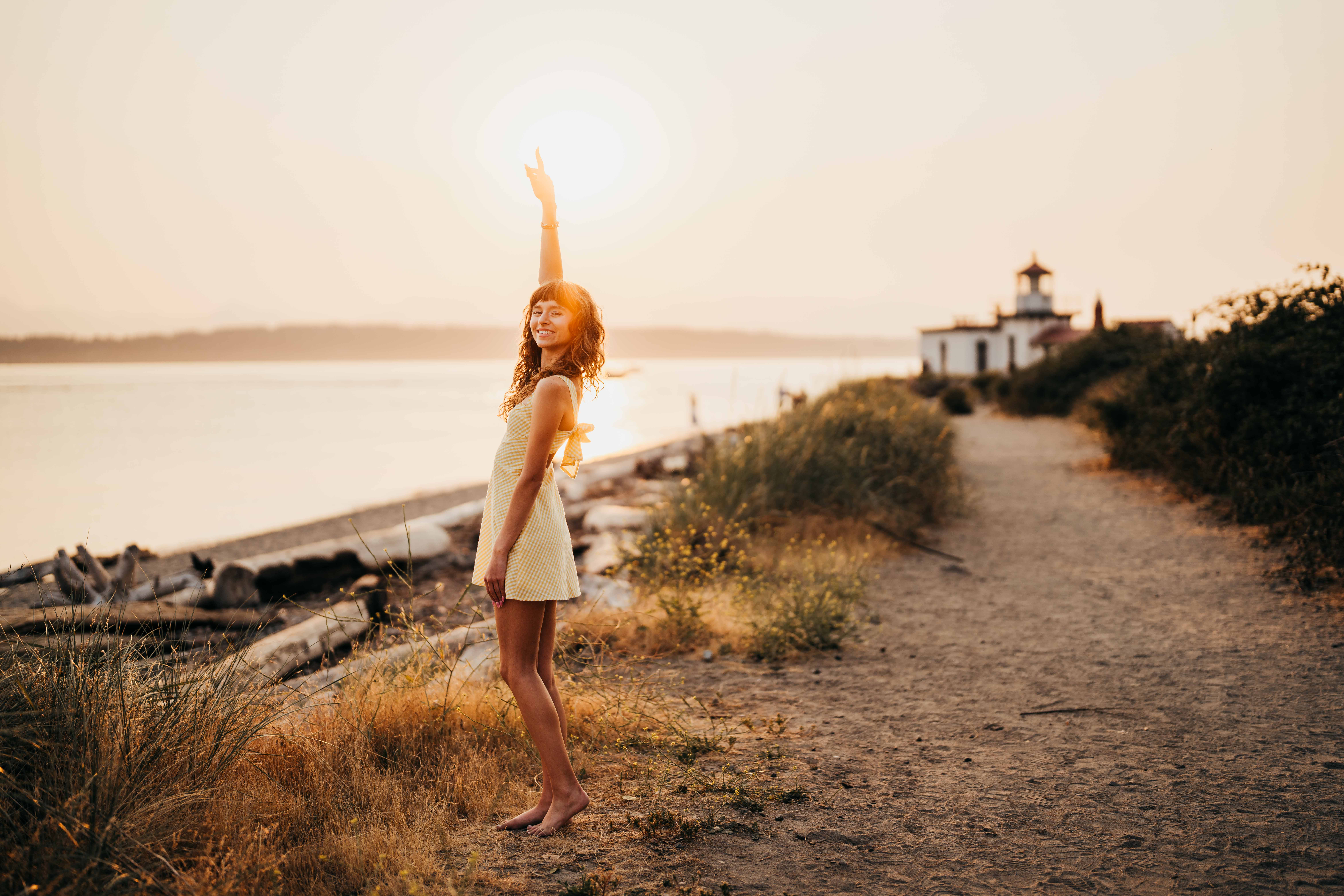 A woman in a yellow dress points to the sky while standing on a waterfront trail at sunset by a lighthouse