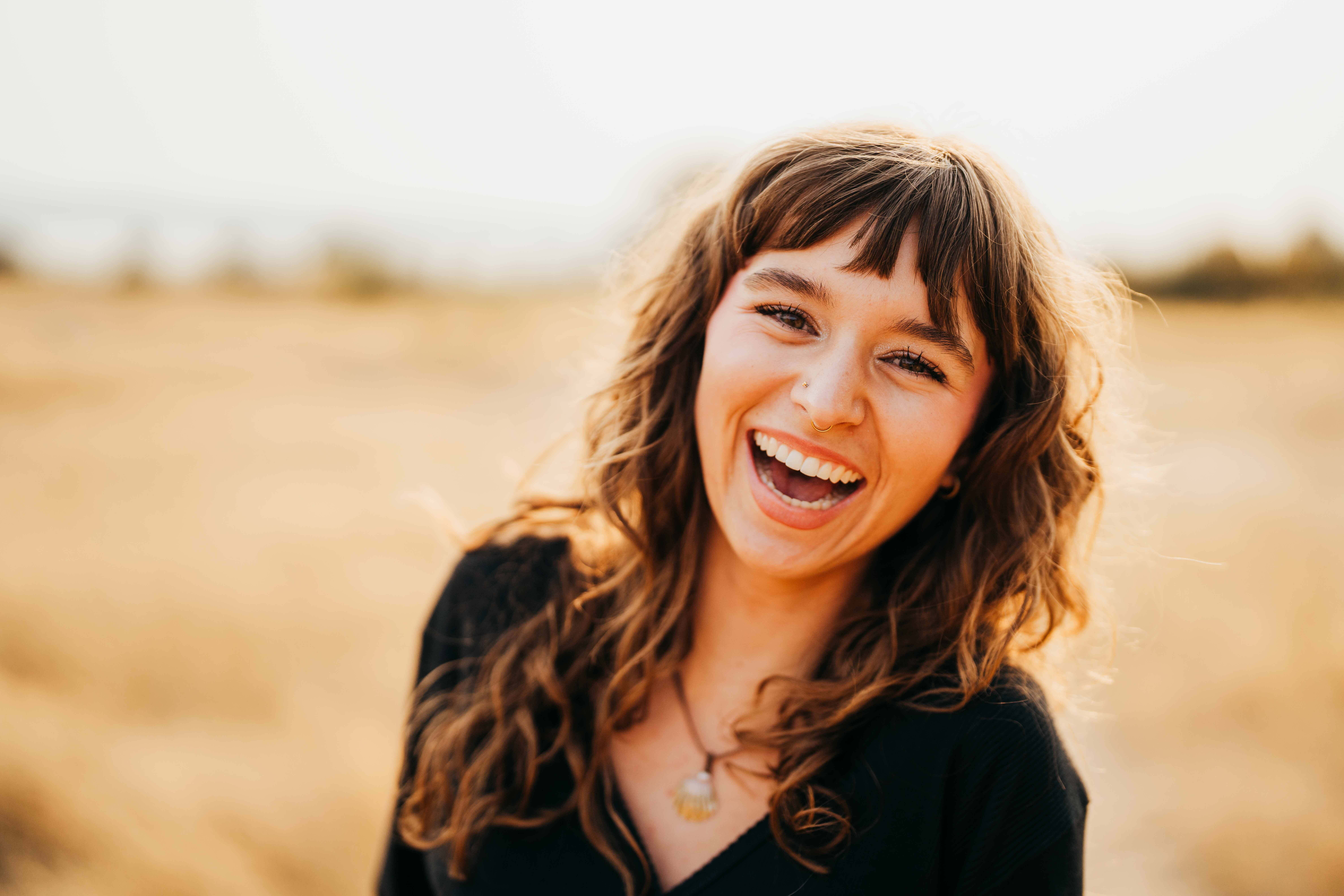 A high school senior in a black shit with a nose ring laughs at sunset in tall golden grass after being a volunteer on mercer island
