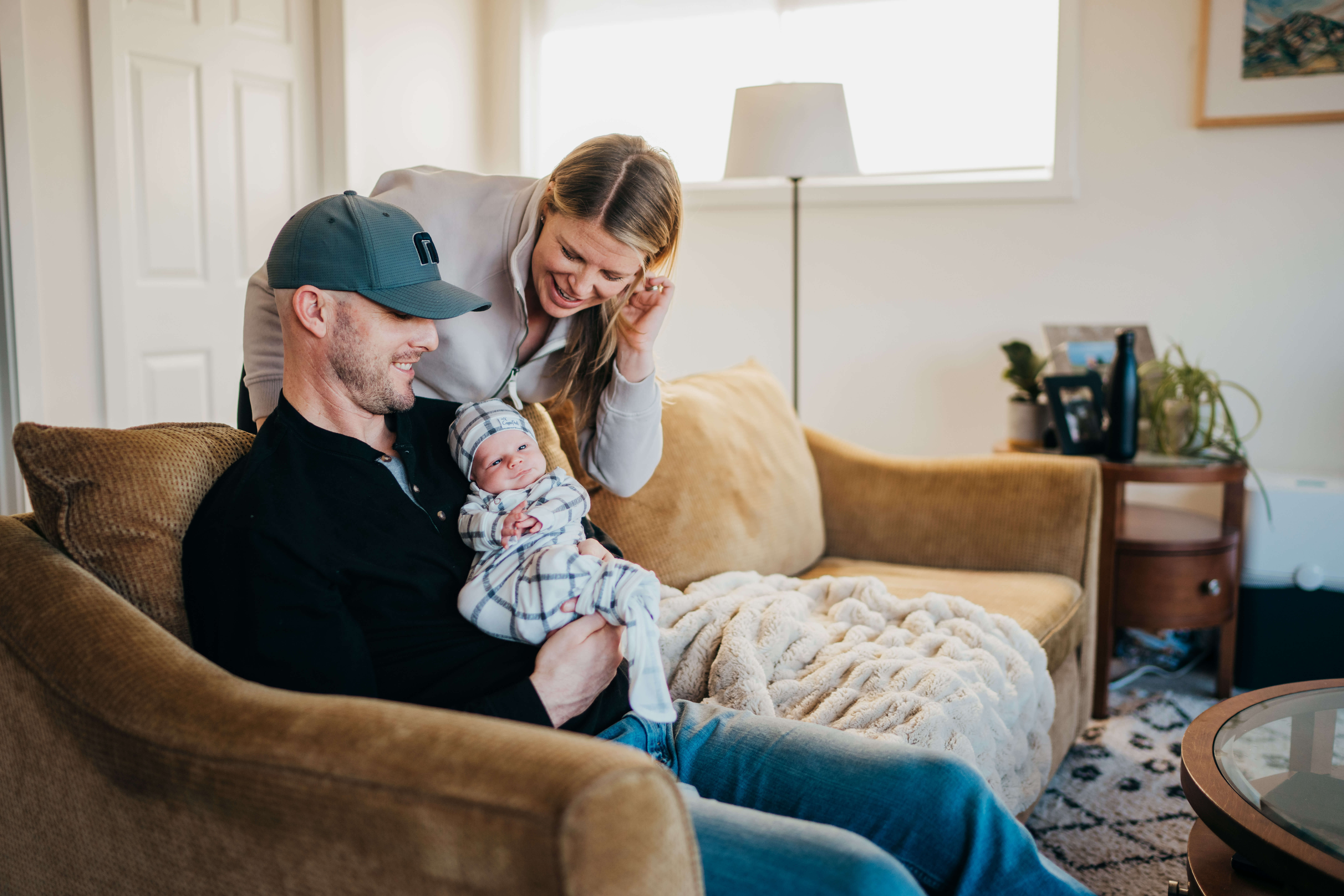 Happy bnew parents snuggle on a couch with their awake newborn in dad's chest in a white stripe swaddle
