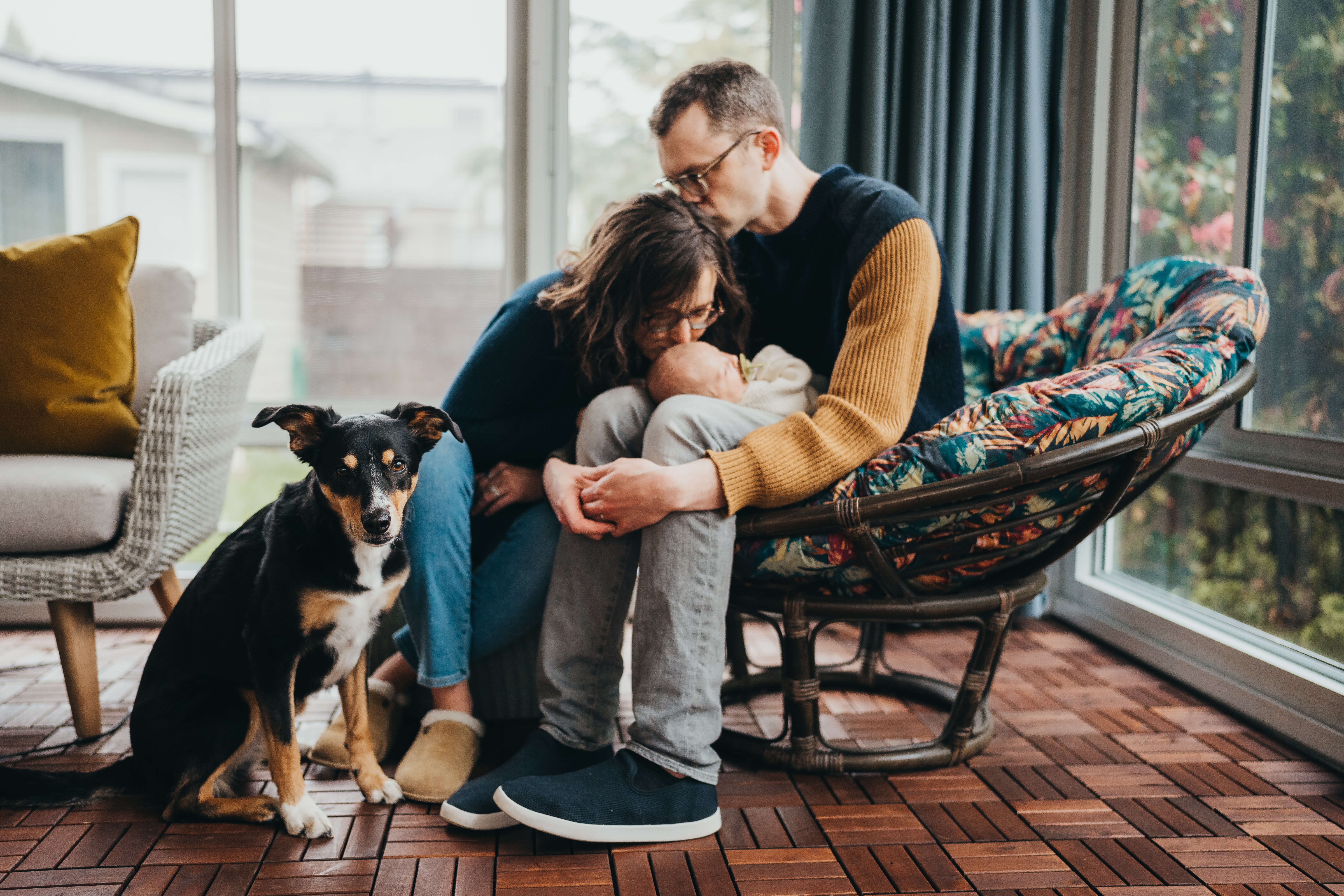 A mom leans in to kiss her newborn in dad's lap in a wicker chair with their dog sitting next to them before finding great date night ideas in seattle