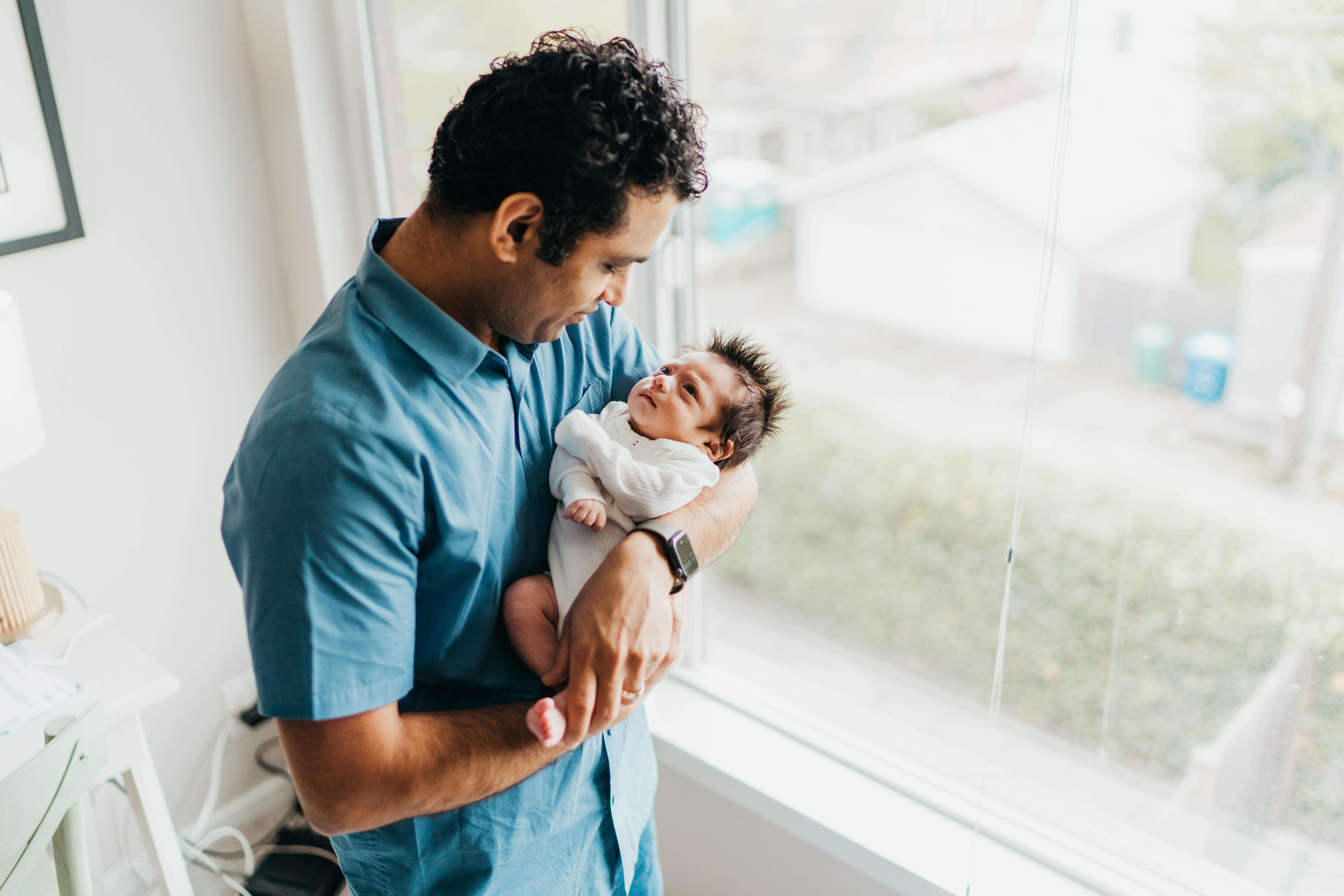 A new dad smiles down to his awake newborn in his arms while standing in a window before a frenectomy in seattle