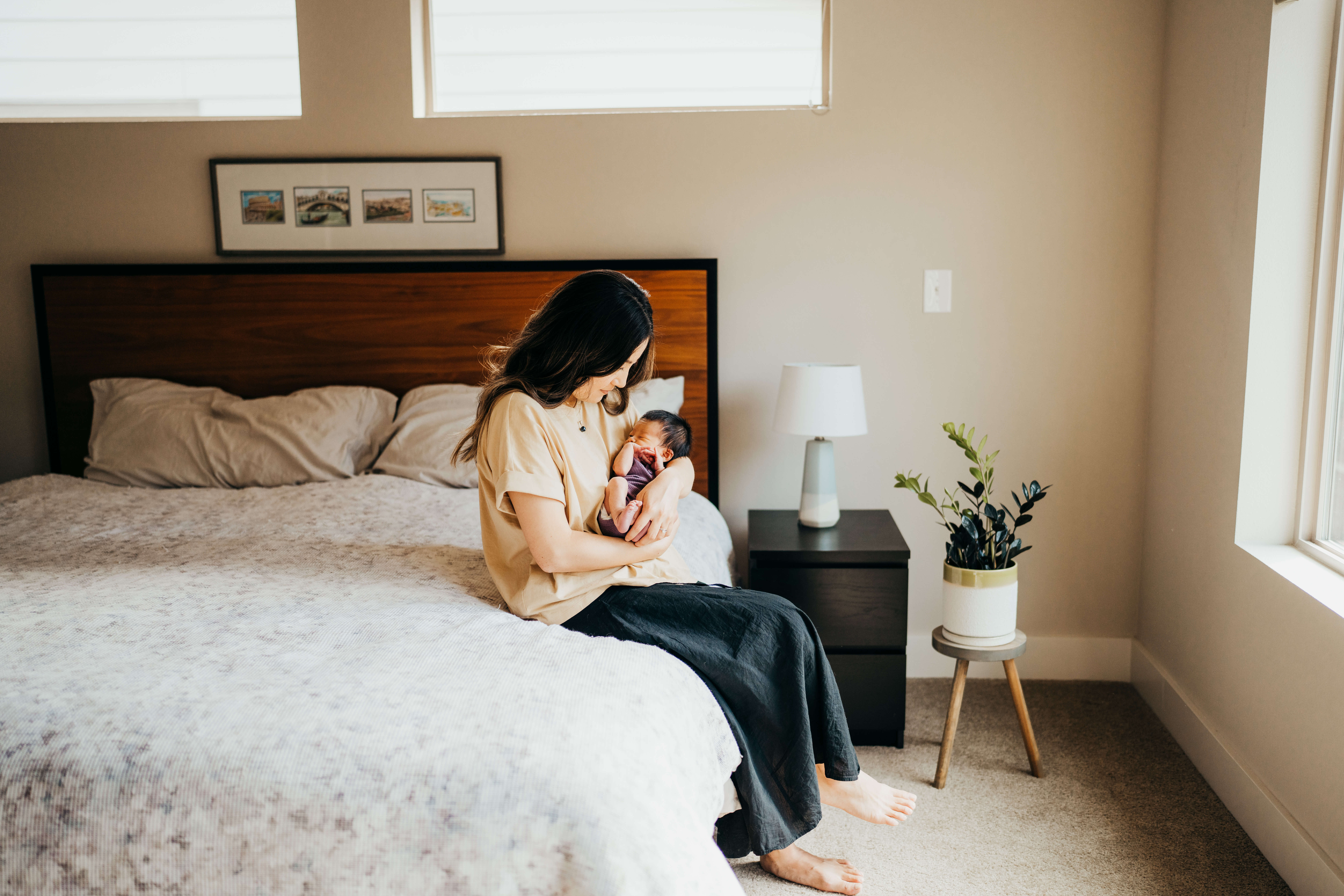 A new mom in a tan blouse and black pants sits on a bed holding her sleeping newborn under a window
