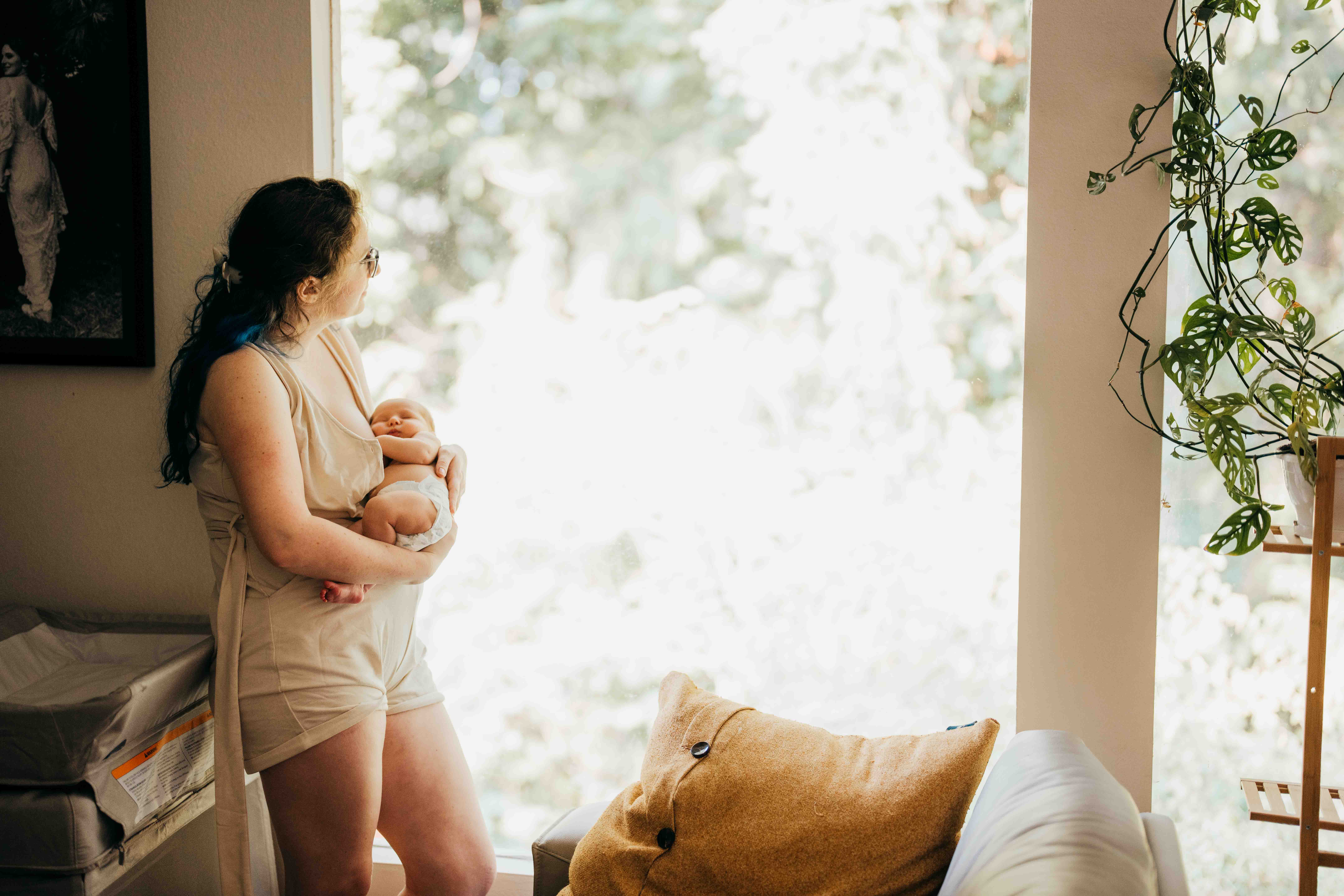 A new mom in a cream romper stands looking out a window cradling her newborn sleeping in her arms before enjoying mother's day brunch in seattle