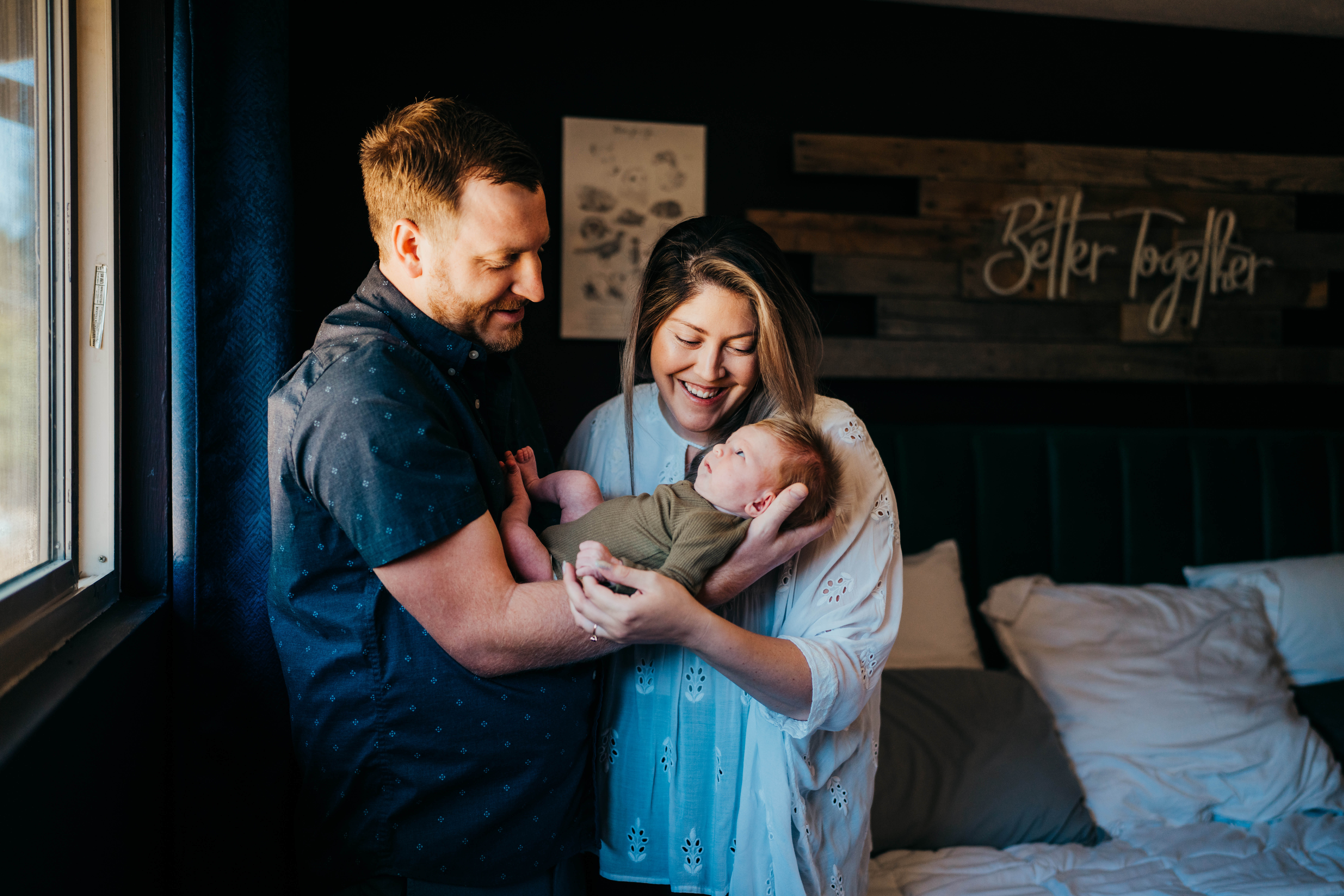 Happy new parents stand in their bedroom in blue smiling down to their awake newborn in their hands in a window before rewarding themselves with romantic restaurants on mercer island