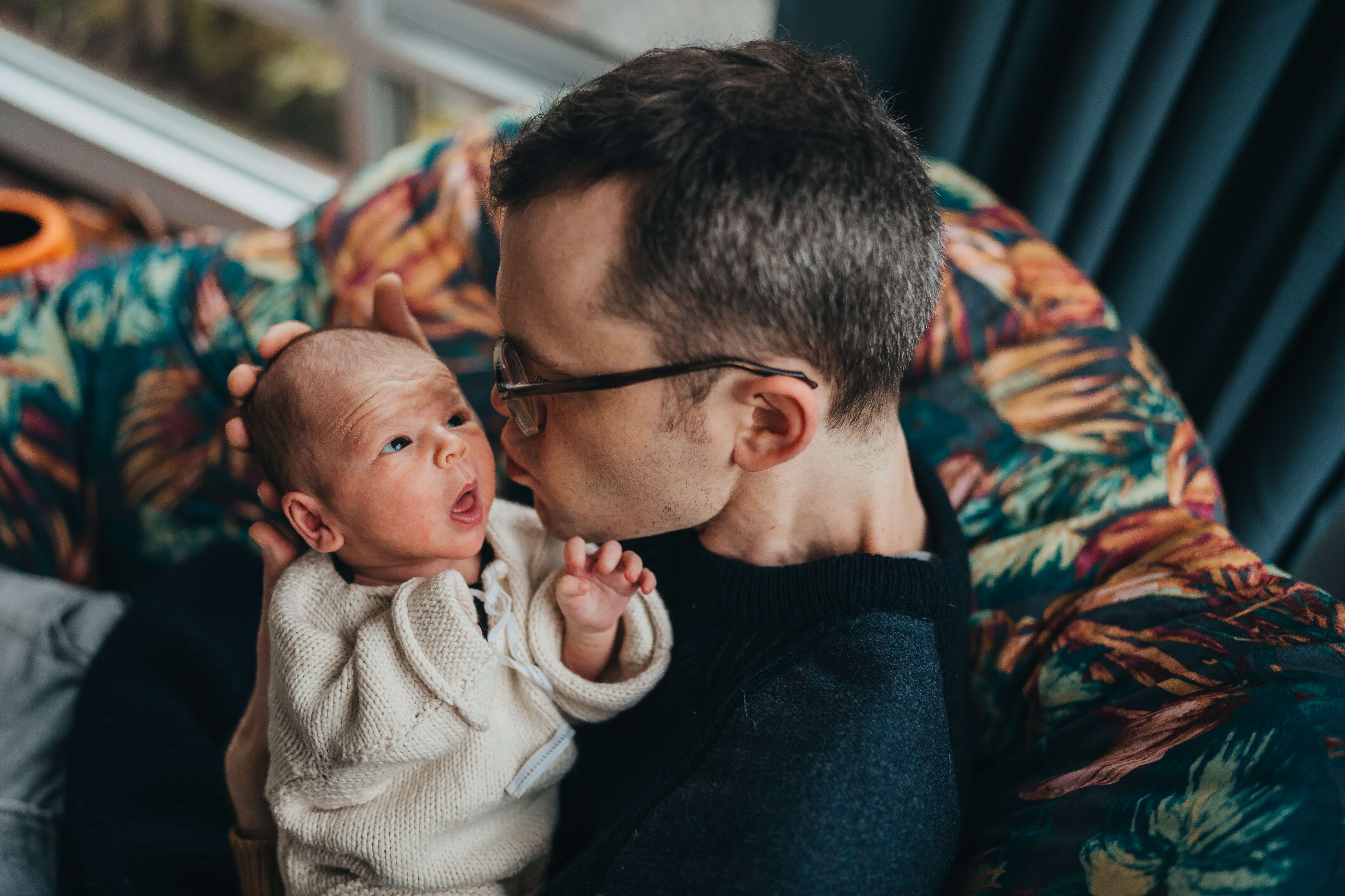 A new dad leans in to kiss his awake newborn wrapped in his arms after visiting aldea home and baby