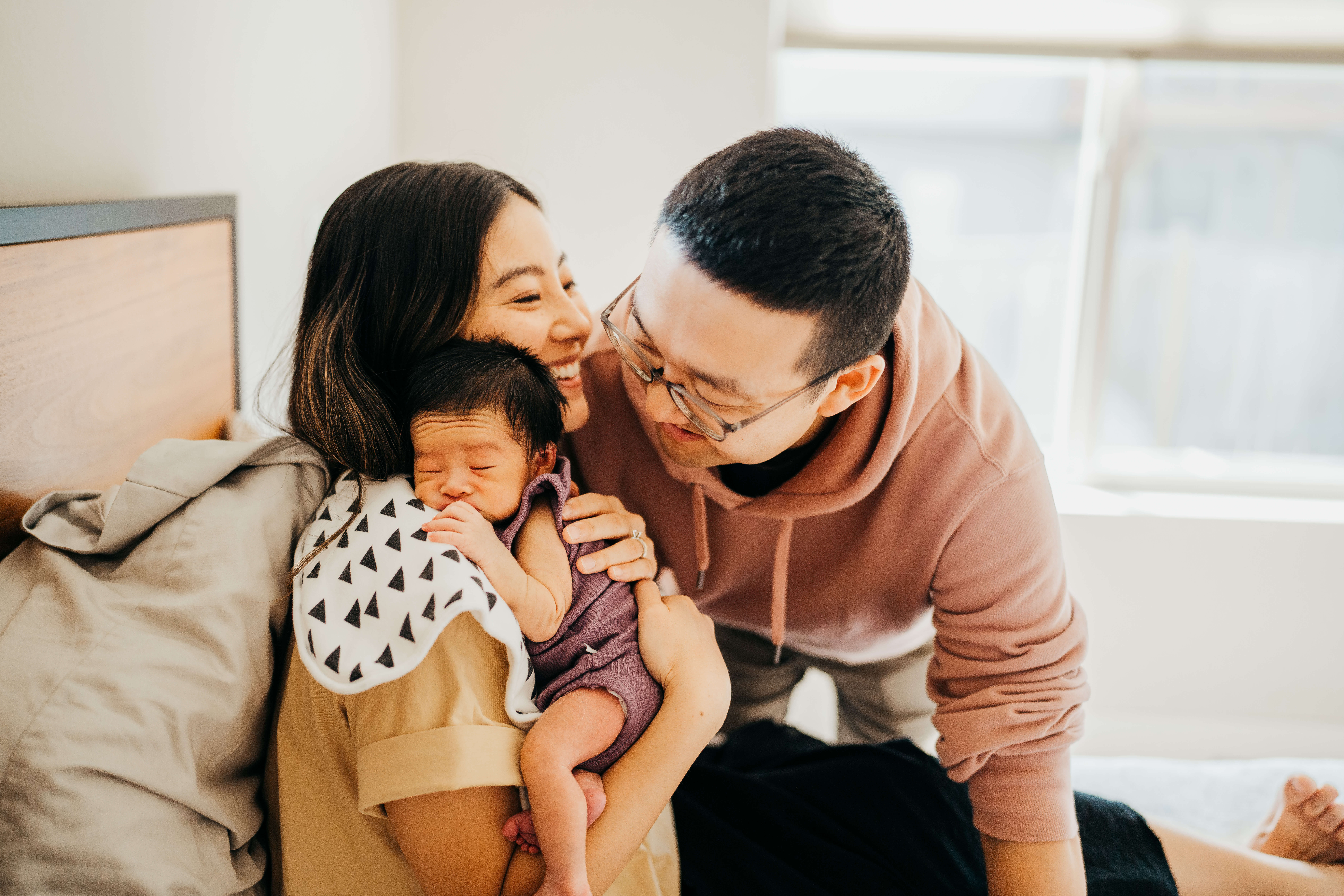 Happy new mom and dad sit on a bed while their newborn sleeps on mom's shoulder after using diaper stork