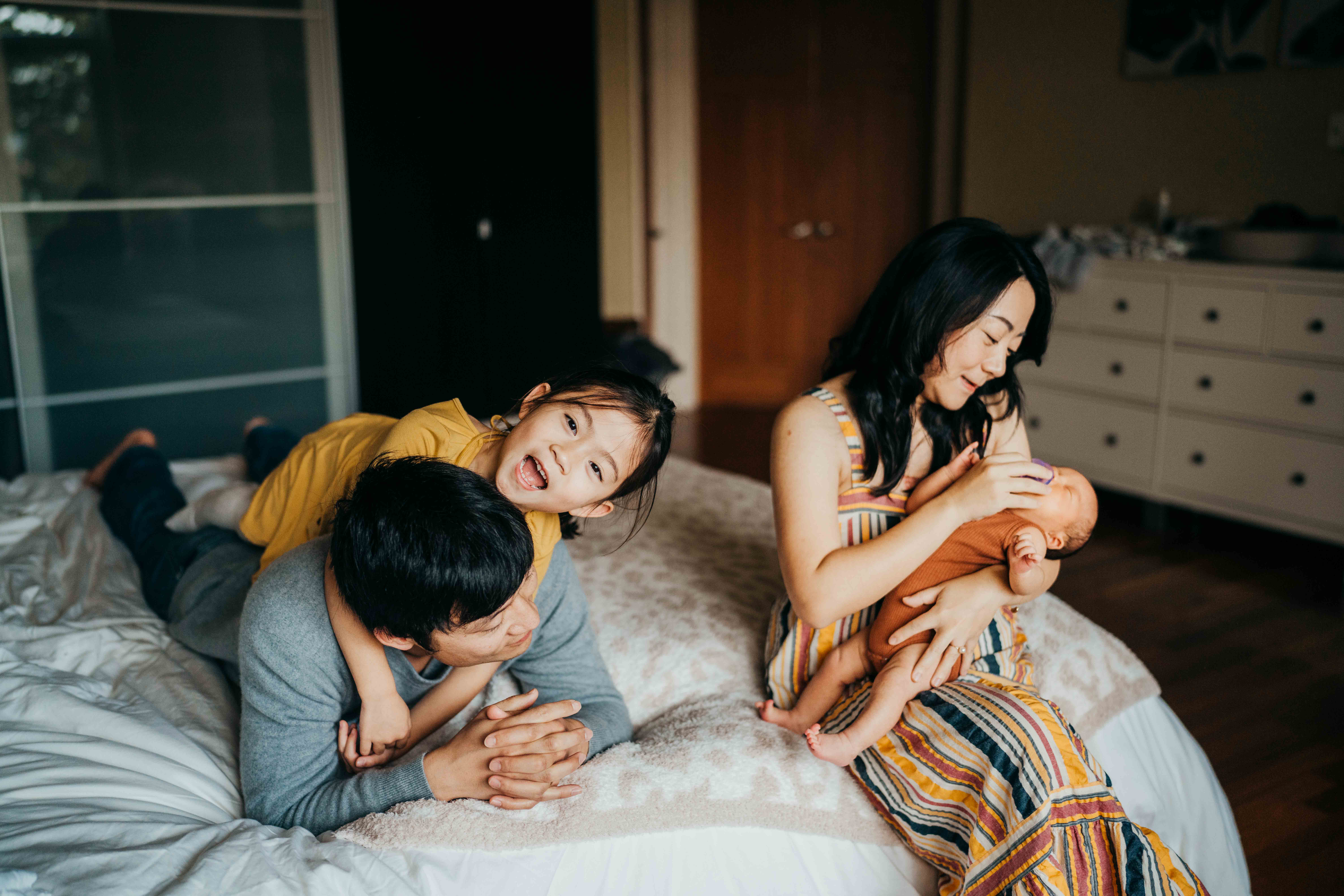A toddler girl climbs on dad on a bed next to mom holding a sleeping newborn in her lap