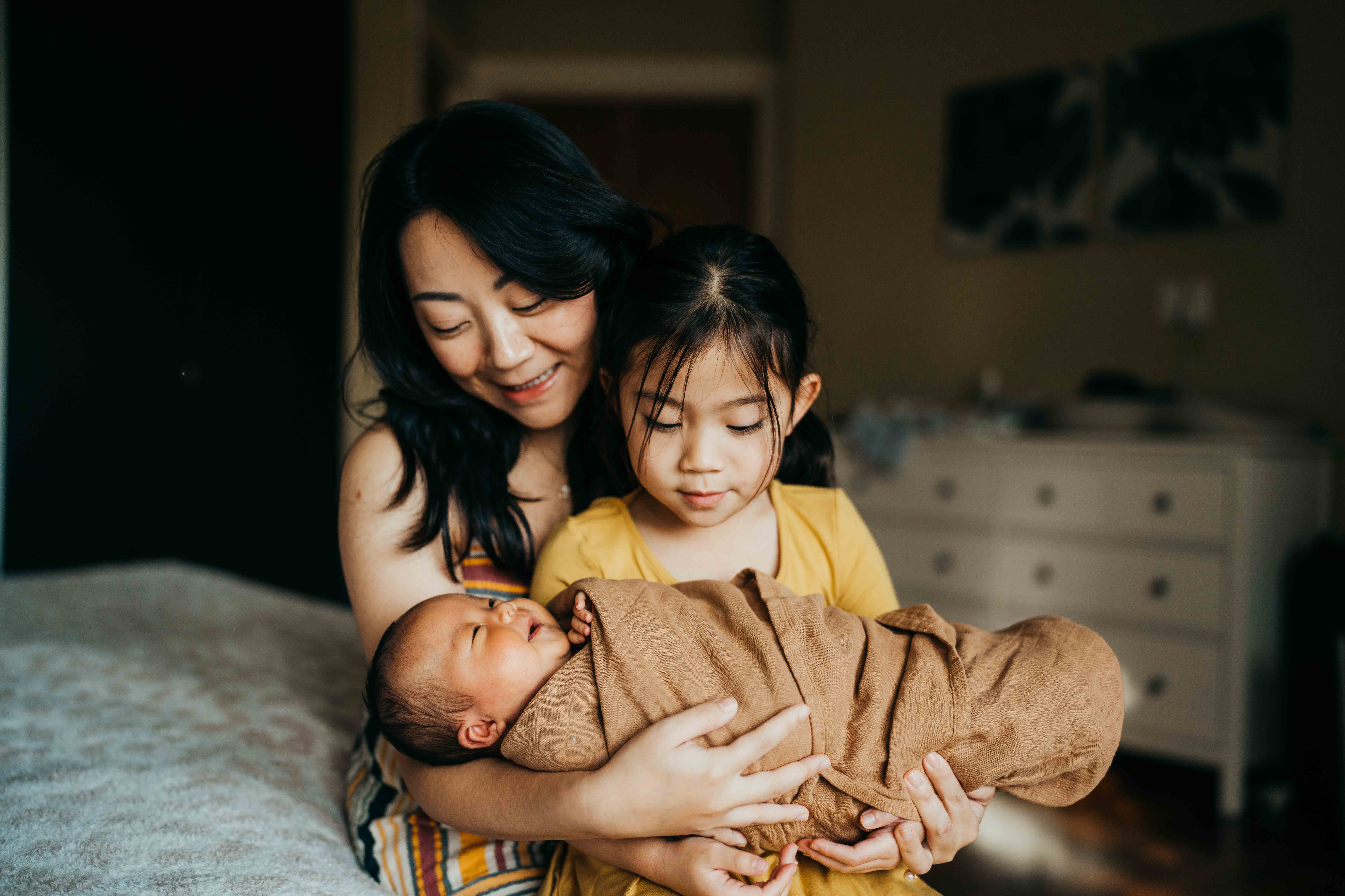 A mother helps her toddler daughter hold her newborn sibling while sitting on a bed thanks to peps seattle