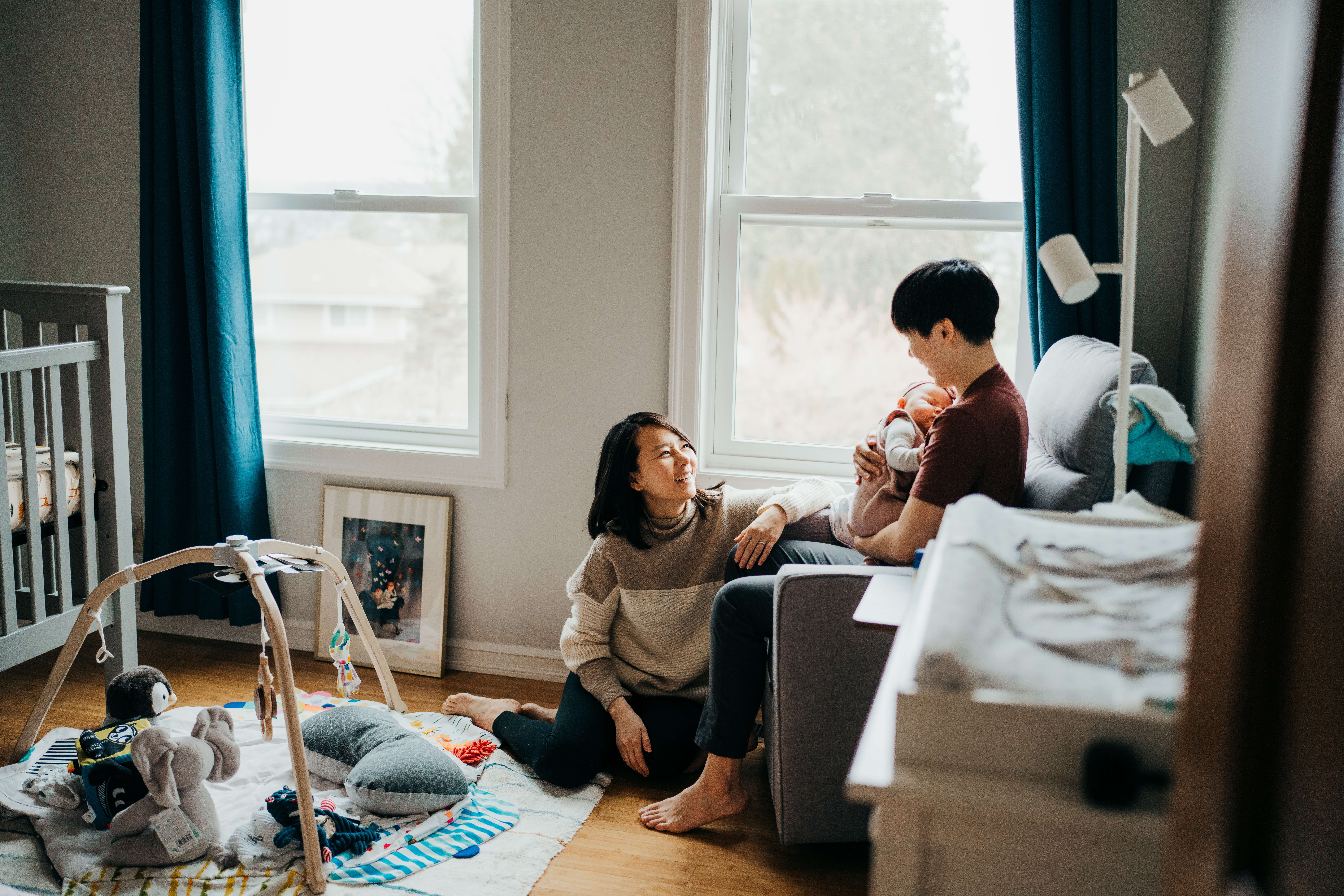 A baby girl sleeps in dad's arms in a nursing chair as mom sits on the floor under a window