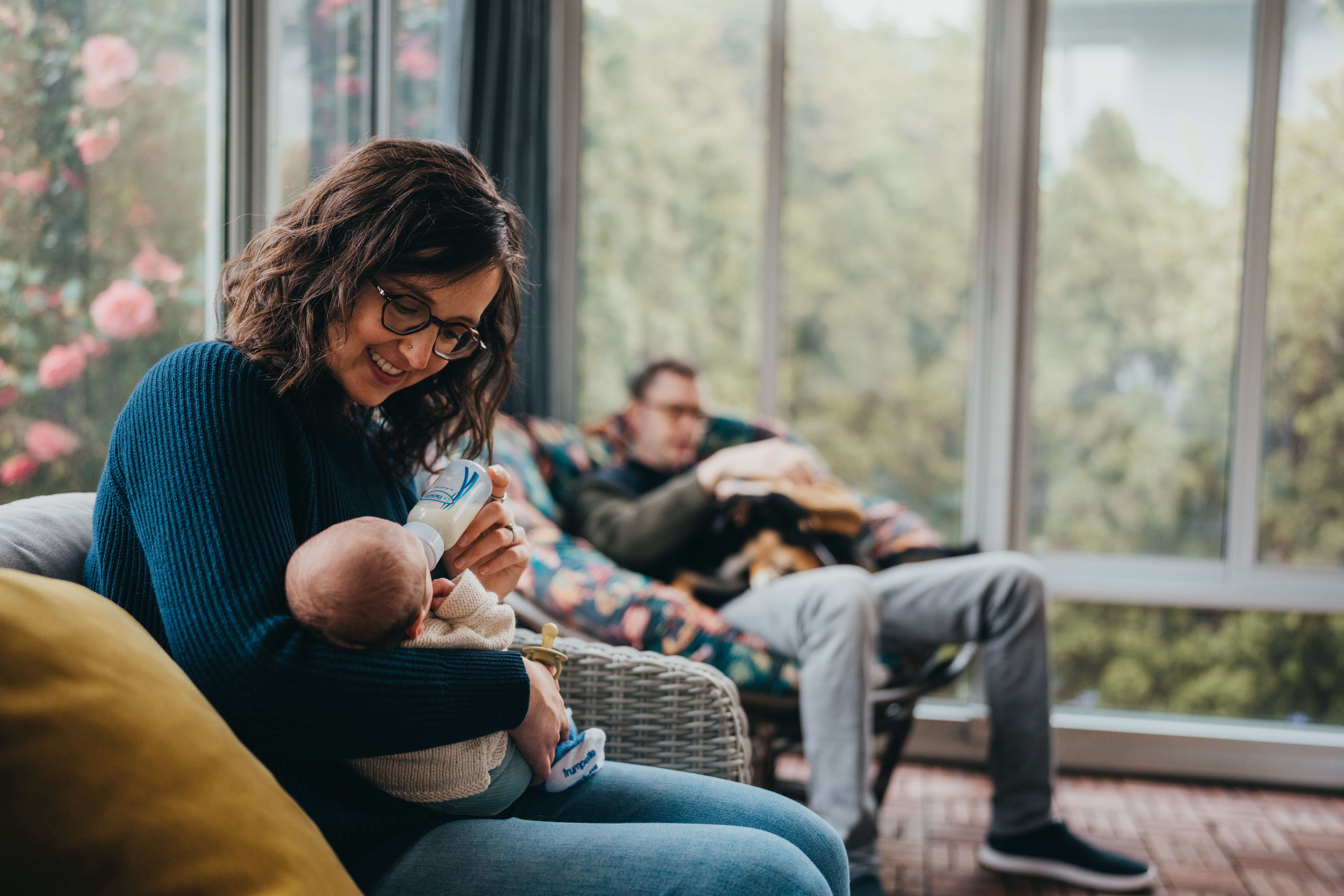 A happy mom feeds her newborn a bottle in a wicker chair in a blue sweater as dad sit behind them after visiting the heybrook seattle