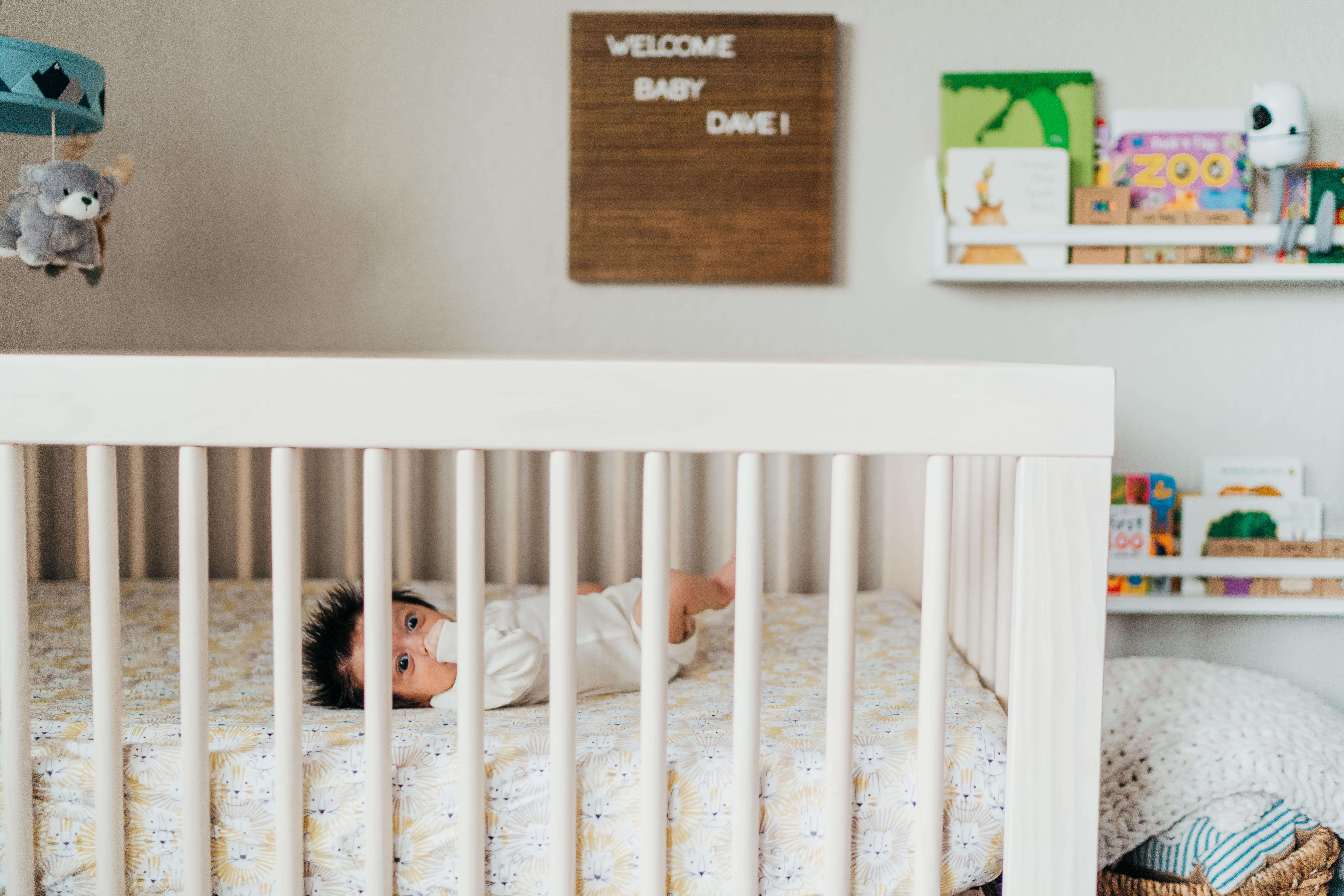 A look at an awake newborn baby in a white onesie in a crib with a hand in his mouth