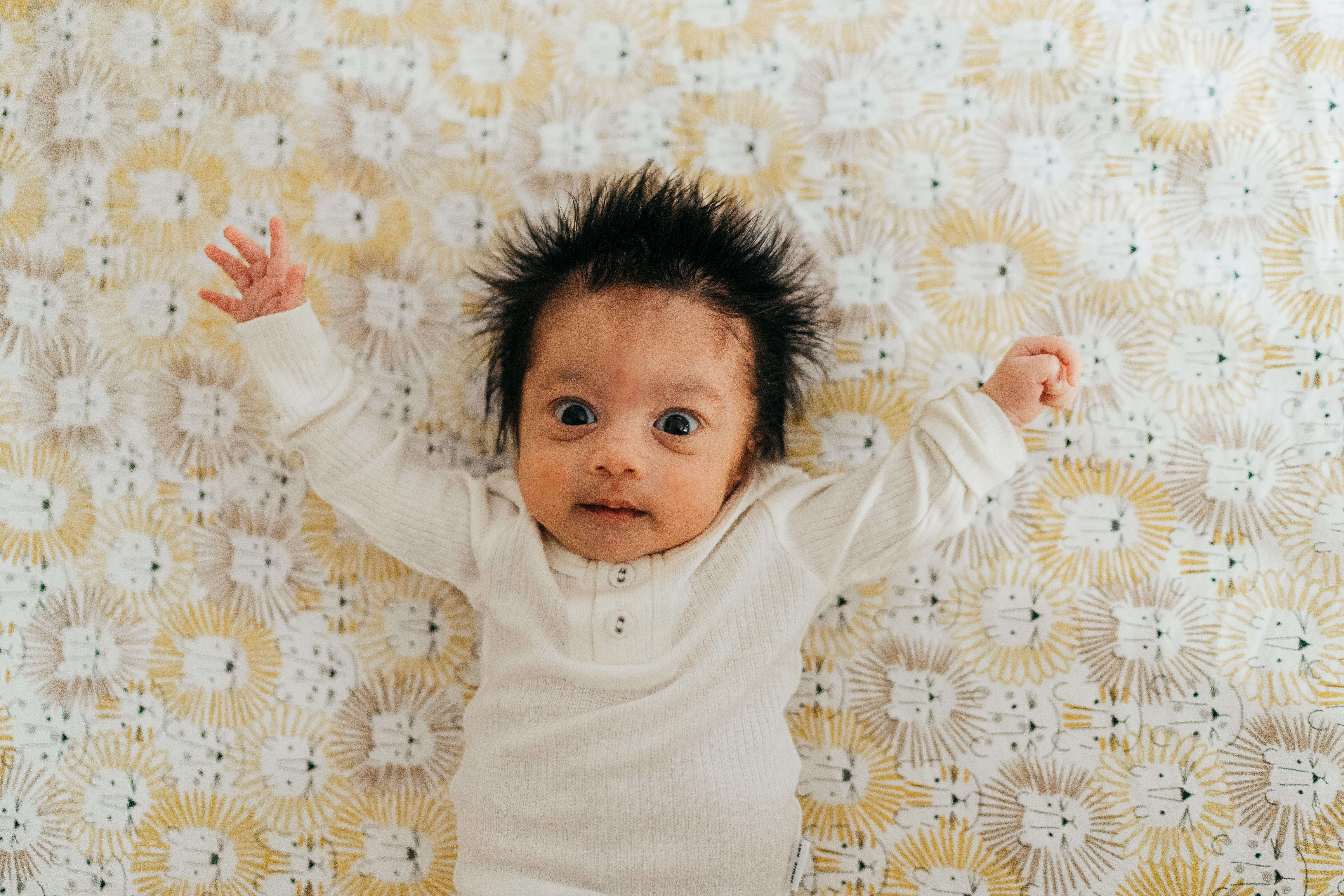 A wide eyed baby lays on a bed with arms wide in a white onesie after visiting two owls seattle